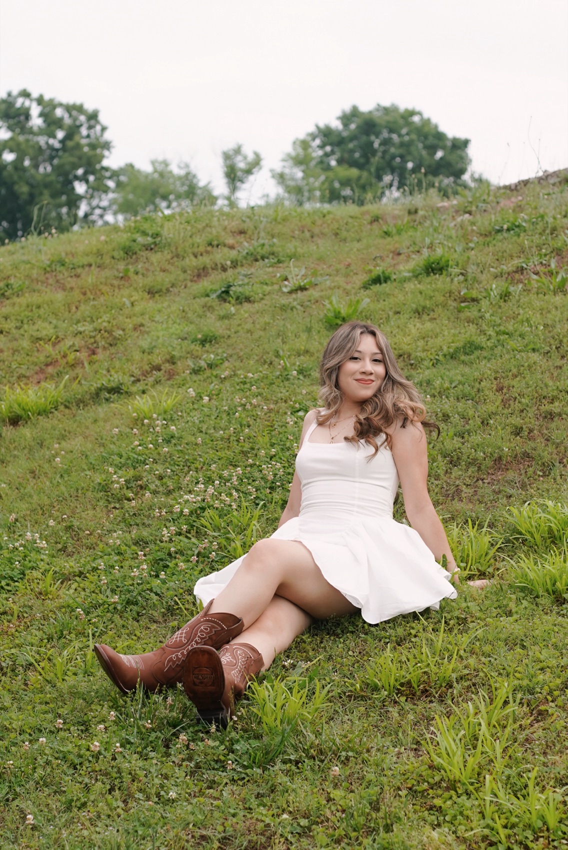 High school senior in a white dress and brown cowboy boots sitting on a green hillside on an overcast afternoon