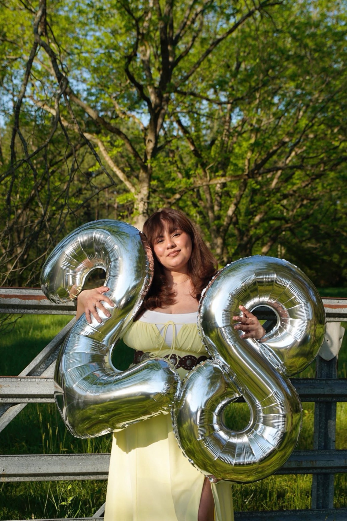 Woman in a pale yellow dress holding silver number twenty-eight balloons in front of a farm gate at golden hour