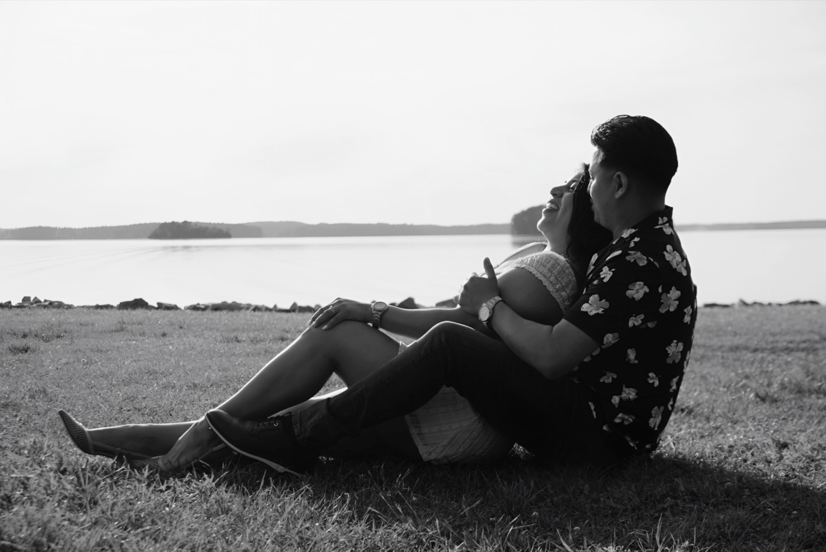 Black and white photograph of a couple sitting together on the grass at the water's edge, backlit by the low sun