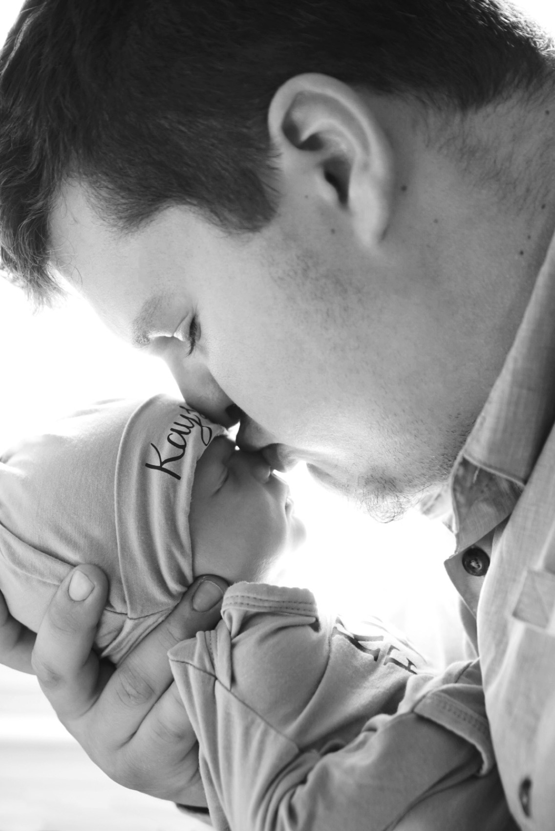 Black and white photograph of a father kissing his newborn's hand, the baby's name embroidered on her hat