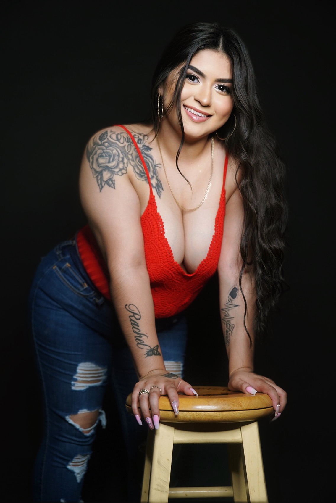 Studio portrait of a woman in a red knit top leaning on a wooden stool against a black backdrop