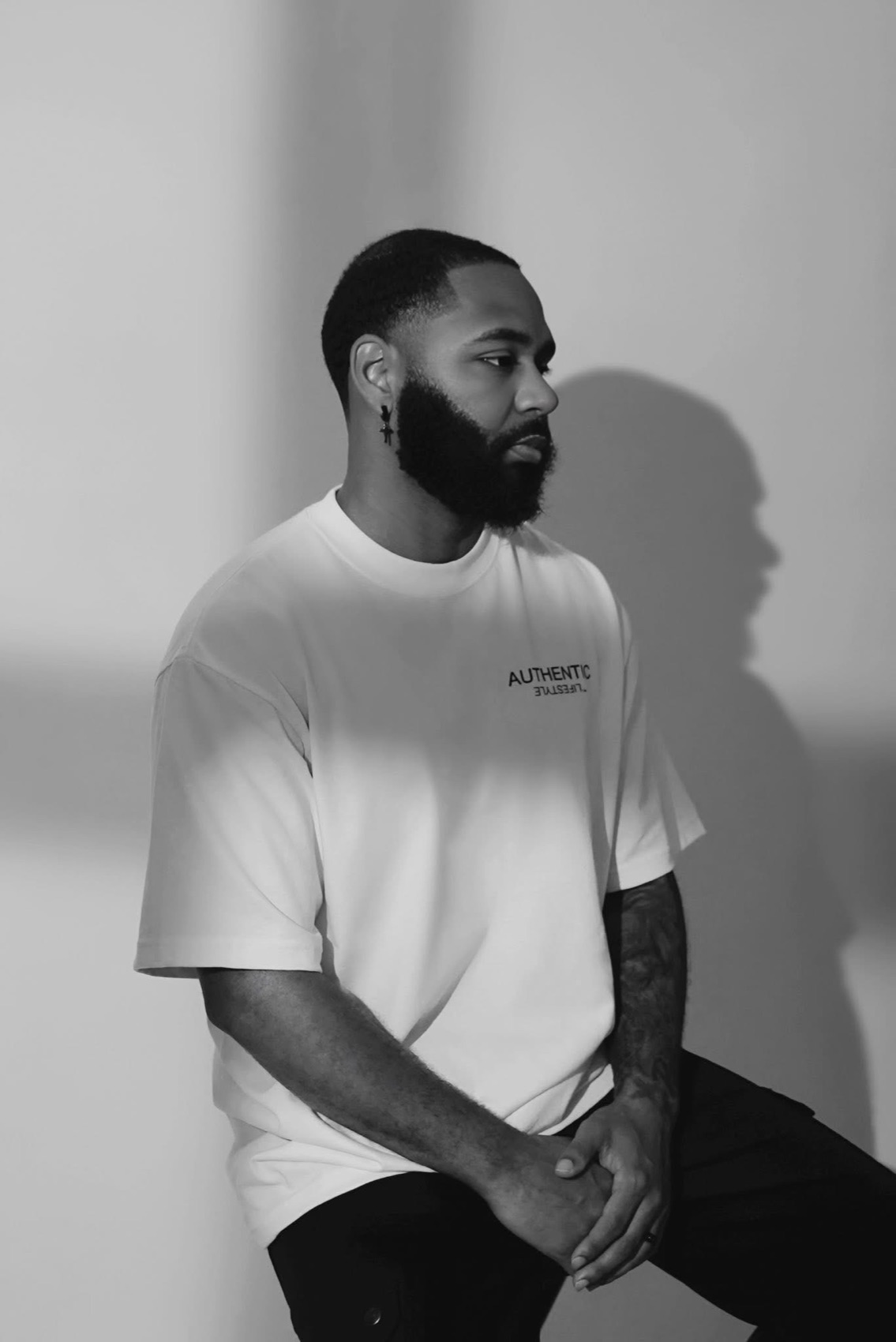 Black and white commercial portrait of a man in an oversized white tee seated against a sunlit studio wall