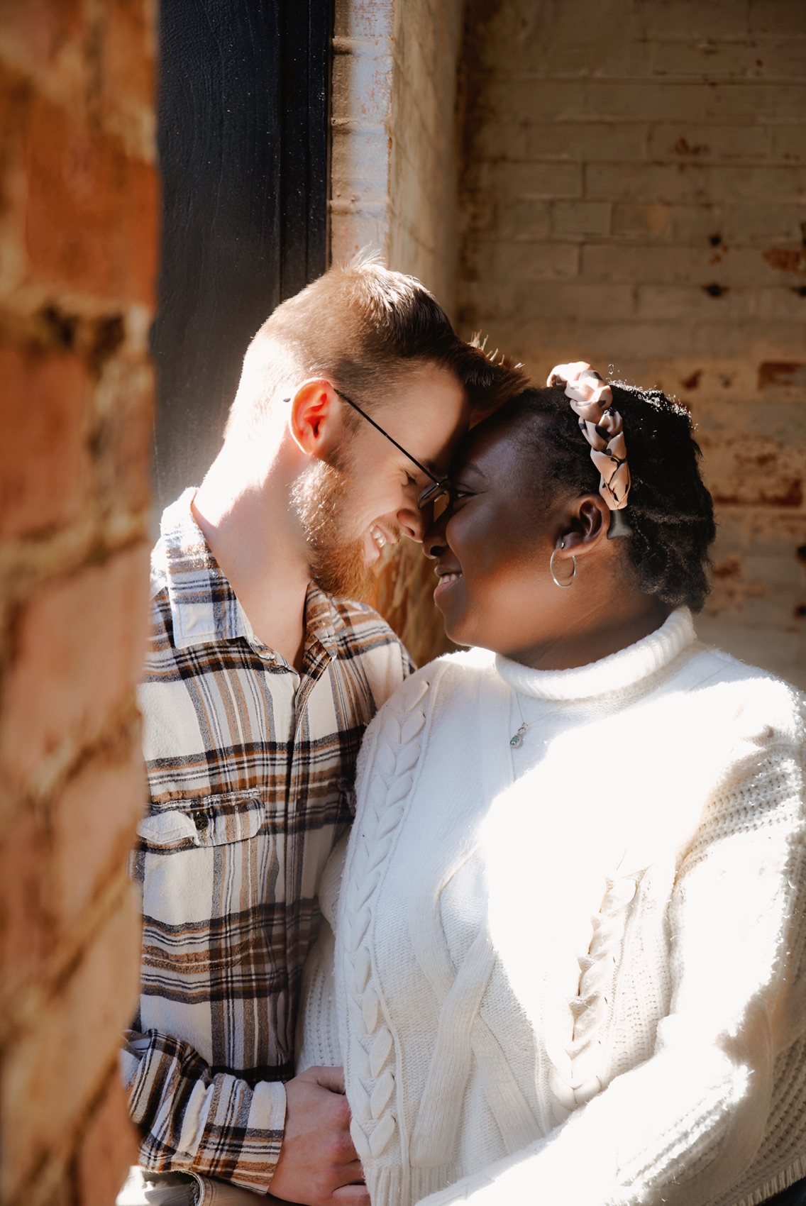 Couple standing forehead to forehead in a shaft of sunlight against an old brick storefront wall