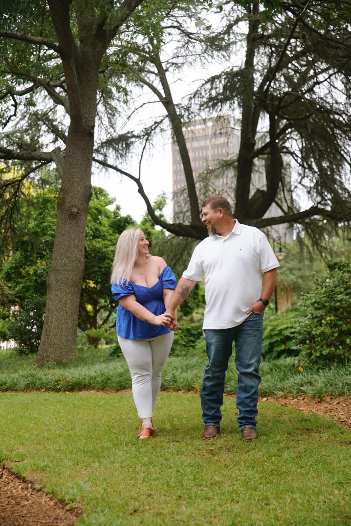 Couple walking hand in hand and laughing beneath old trees in a downtown park, the city skyline behind them