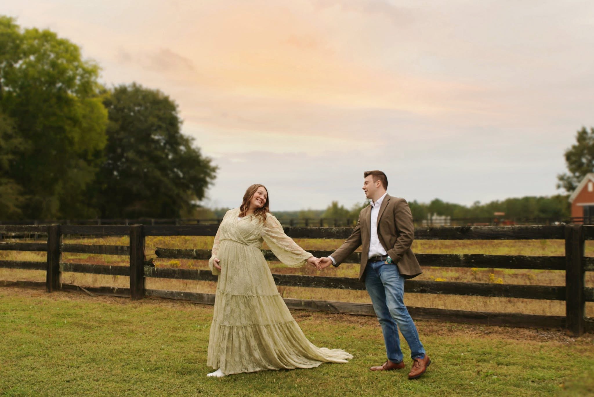A couple holding hands and laughing beside a wooden fence in a pasture at dusk under a pastel sky, photographed by Rendon Photography