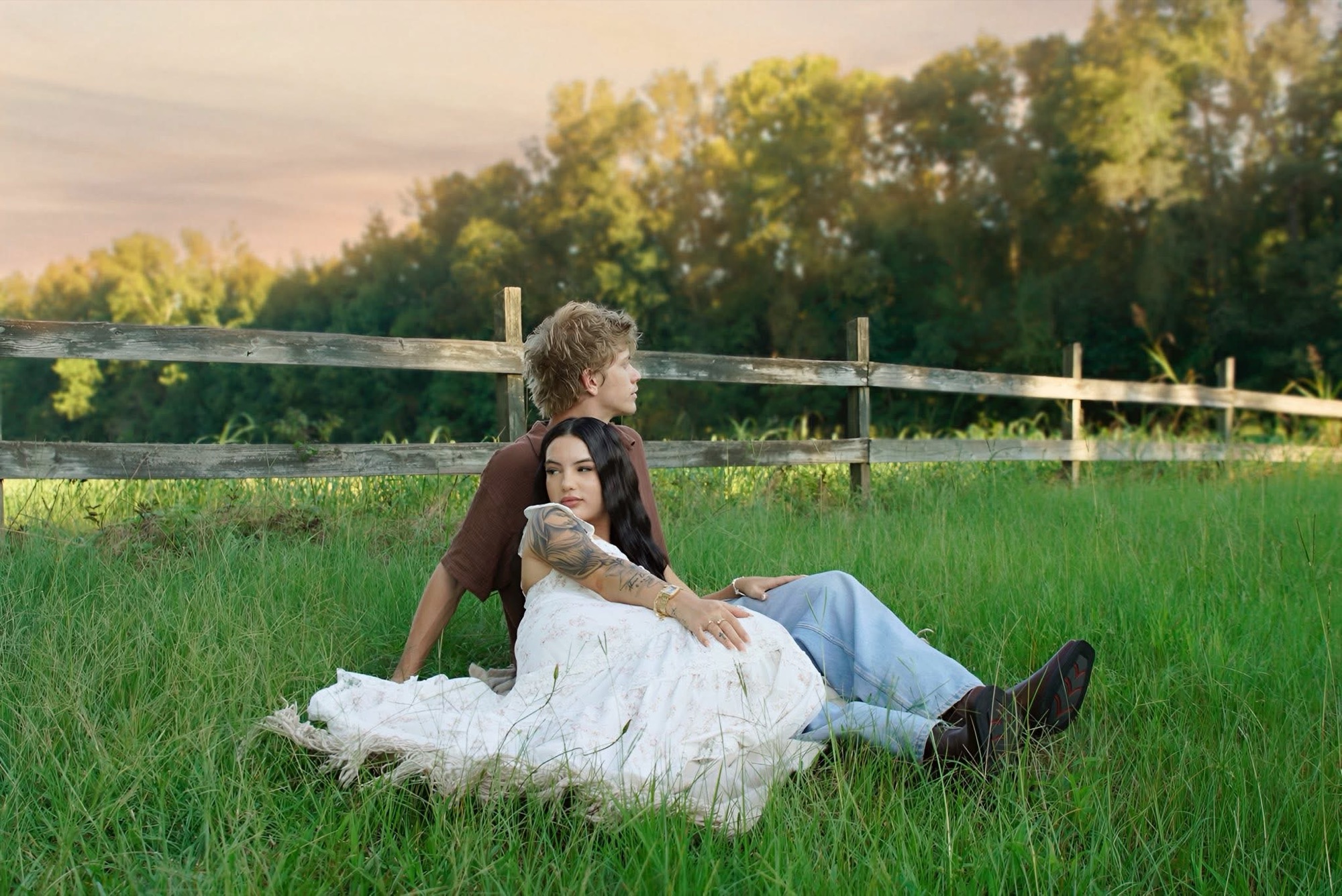 A couple resting together in tall summer grass beside a wooden fence line at sunset, photographed by Rendon Photography near Newberry, South Carolina